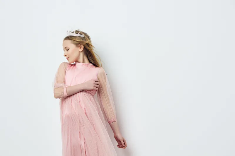 Young girl in pink dress gently posing with crown, exuding joy and innocence against a minimalist white background, perfect for fashion and children s photography