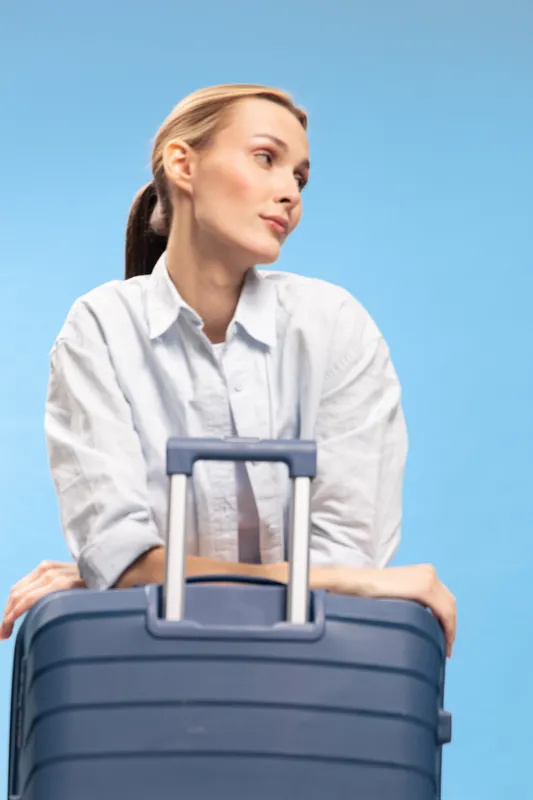 Young woman in white shirt leans on blue suitcase, looking sideways with thoughtful expression on blue background. Concept for travel, waiting, and journey preparation.