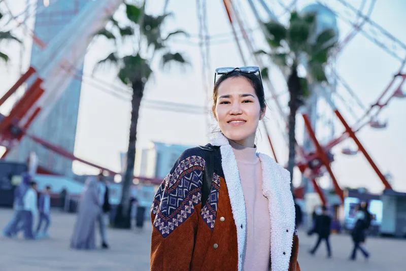 Woman wearing a patterned brown jacket and pink turtleneck stands in a sunny urban street with palm trees and glass buildings in the background, showcasing contemporary outdoor fashion and city vibe