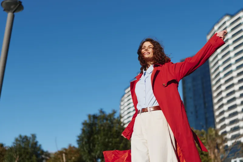 A joyful woman with curly hair in a red coat and white shirt raises her arms outdoors, smiling against a clear blue sky and a modern city backdrop as warm afternoon light highlights her pose.