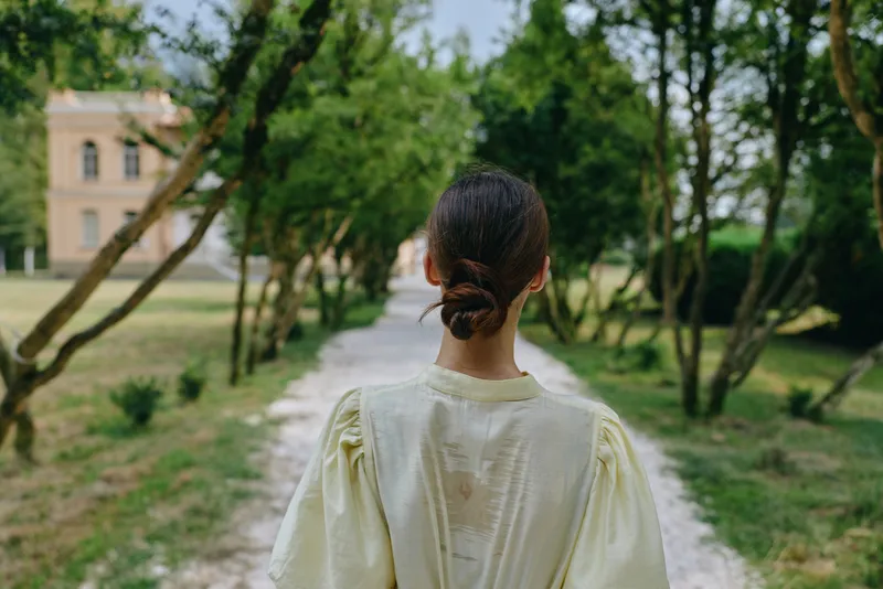A woman stands with her back to the camera along a gravel path in a lush park, wearing a pale dress, surrounded by green trees and open space.