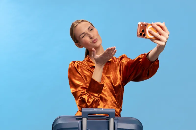 Young woman in orange blouse taking selfie with smartphone while standing behind suitcase on blue background. Concept of travel, vacation, and social media communication.