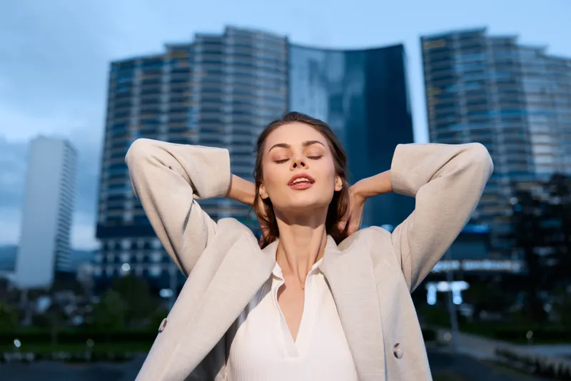 Confident businesswoman enjoying a moment of relaxation outdoors, wearing a light blazer, against a modern city skyline background.