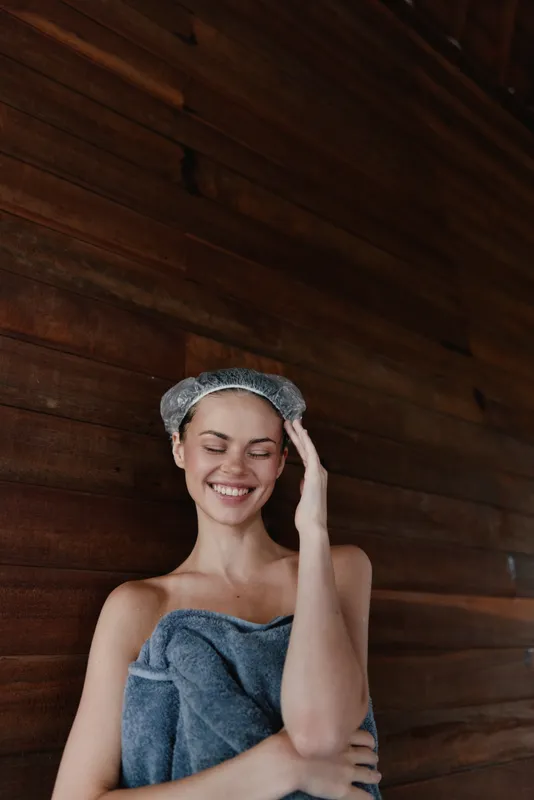 Smiling woman wrapped in towel and wearing shower cap stands happily against rustic wooden wall, enjoying spa moment with relaxed and joyful expression, cozy indoor setting.
