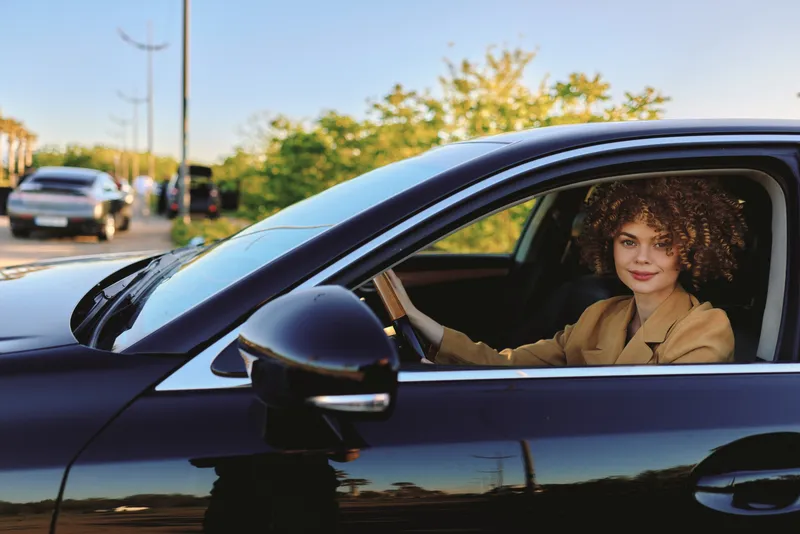Cheerful woman driving a car, curly hair, sunlit street, retro film color, casual blazer, relaxed expression, urban mood, daytime portrait, candid motion