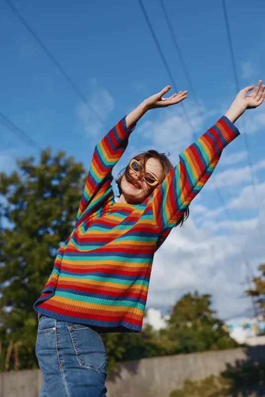 Woman in a rainbow sweater wearing sunglasses and smiling outdoors, joyful portrait with arms raised, casual outfit and blue sky background, carefree youth and vibrant fashion.