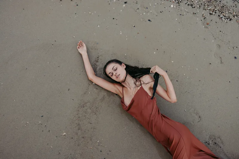 A woman lies on sandy ground in a flowing red dress, arms stretched above, eyes closed, creating a dramatic moment of stillness, grace, and soft natural light.