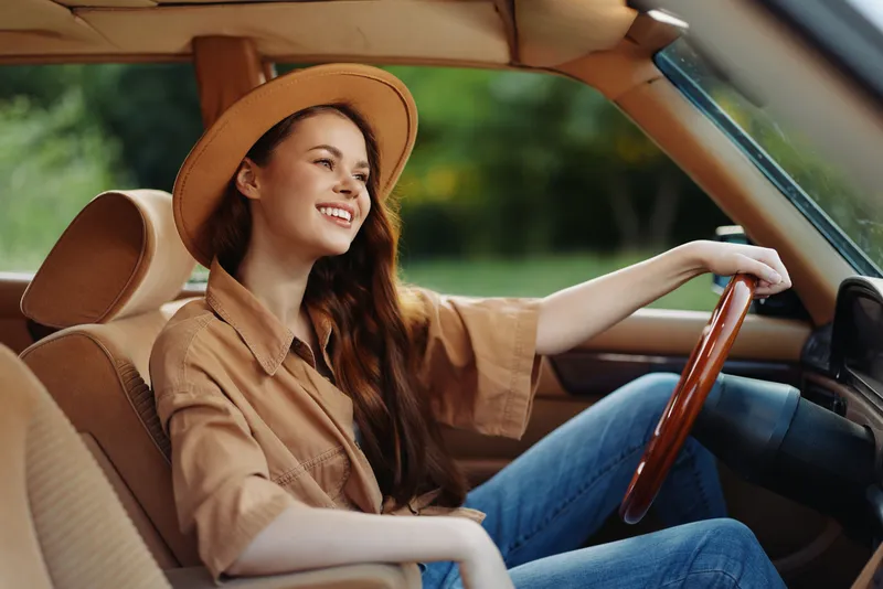 Young woman with long hair wearing a hat, smiling joyfully while sitting in a vintage car, embodying a carefree summer vibe in a green outdoor setting.