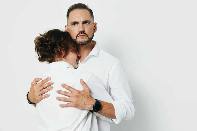 A professional man in a white shirt embraces a colleague, signaling support and calm under pressure. The isolated white background highlights emotion, focus, and leadership in a corporate setting.