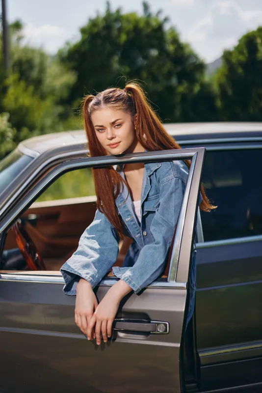 Young woman with ponytails in denim jacket leaning out of a vintage car, smiling in a lush green outdoor setting, conveying a sense of freedom and adventure.