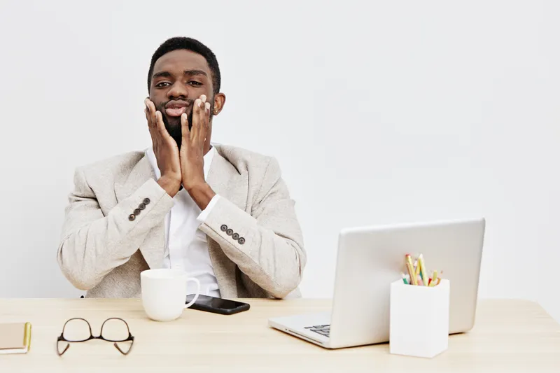 Frustrated young African American man in formal blazer sitting at desk with laptop, coffee mug, glasses, and smartphone, illustrating workplace stress and anxiety, indoor office environment, neutral b