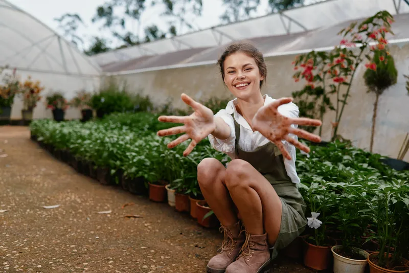 Cheerful young woman in overalls and boots showing her hands in a greenhouse, surrounded by green plants and flowers, representing gardening and nature appreciation