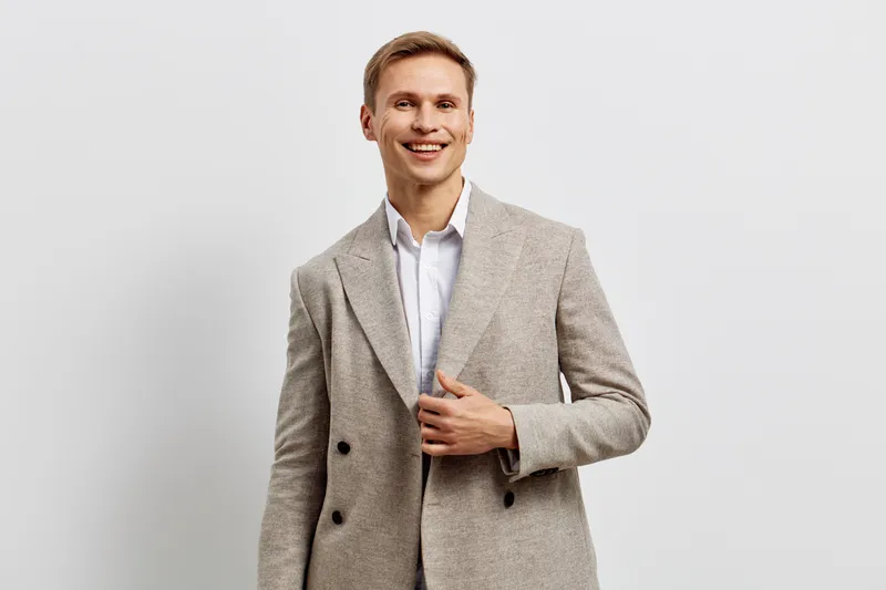 Confident smiling young man in light gray suit with white shirt, casual fashion, standing against plain white background, studio portrait, business style, professional appearance, happy expression, le