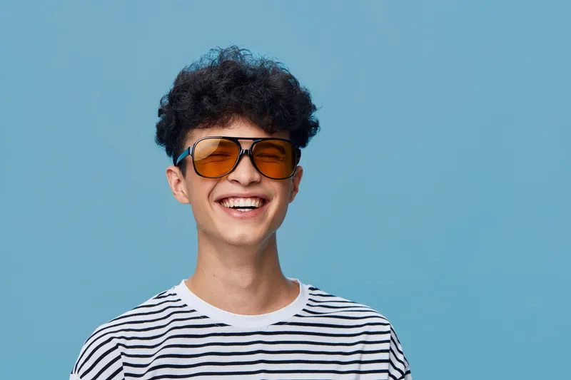 smiling man wearing orange sunglasses and striped t-shirt with curly hair on blue background, bright cheerful expression, casual style portrait