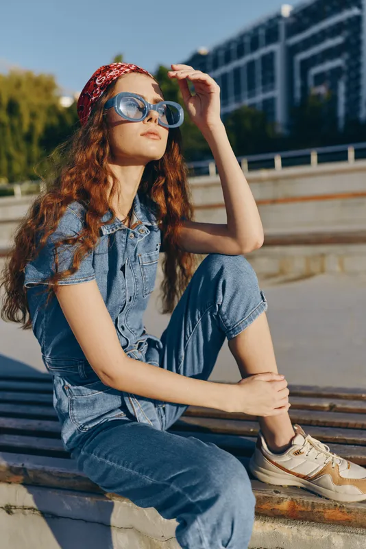 Lifestyle woman in modern boho-western denim street style sits casually outdoors wearing sunglasses and a red bandana, with warm film color tones enhancing the relaxed mood.