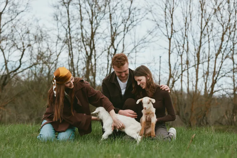 Three young adults enjoy a casual outdoor moment with two baby goats in a grassy field during early spring, capturing a warm and natural lifestyle scene.