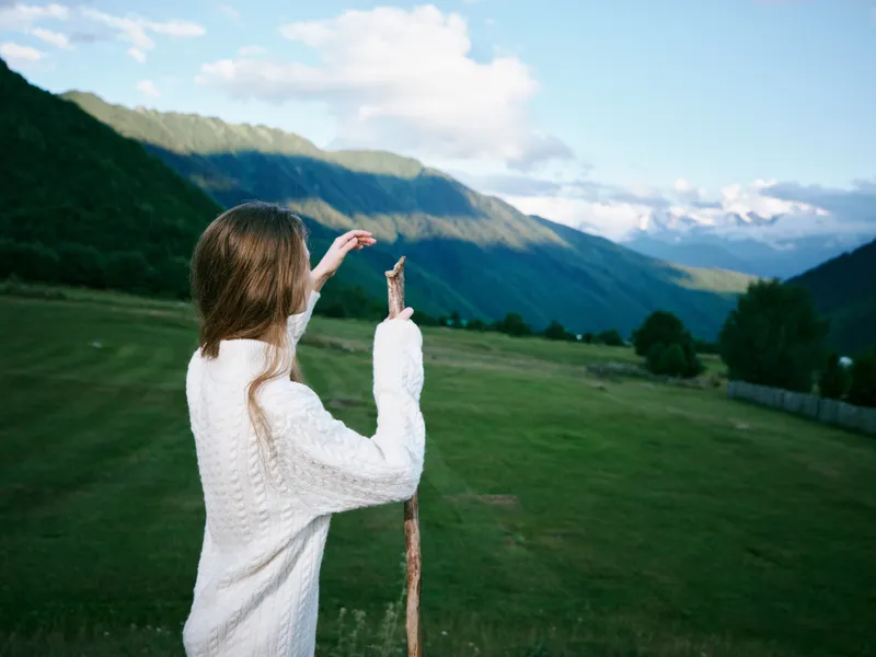 Woman in a knit sweater stands in a field, holding a staff and gazing toward distant mountains, capturing a quiet outdoor landscape under a clear sky and peaceful countryside