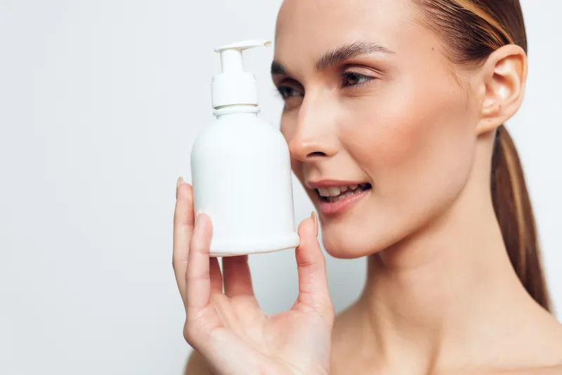 Young woman with clear skin holds white pump bottle close to face, looks at product with slight smile, isolated on white background, skincare and beauty concept.
