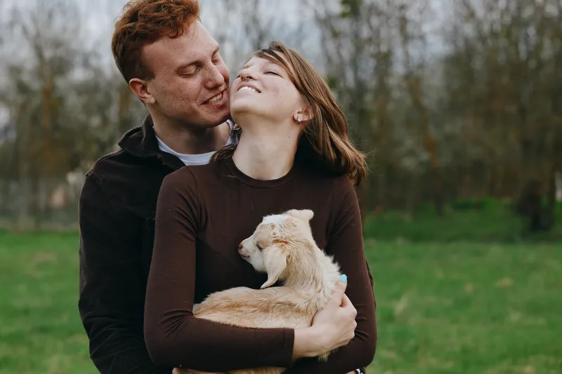 Young couple embraces outdoors while woman holds a baby goat in her arms, sharing a tender moment in a natural green setting during a casual lifestyle shoot.