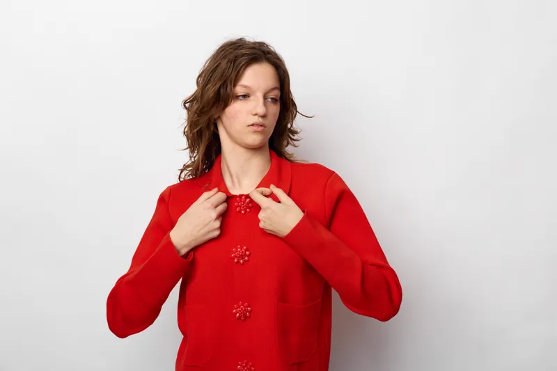 Young woman in red sweater looks down with uncertain expression, adjusting her collar. Isolated on white background. Concept of doubt, hesitation, and casual fashion.