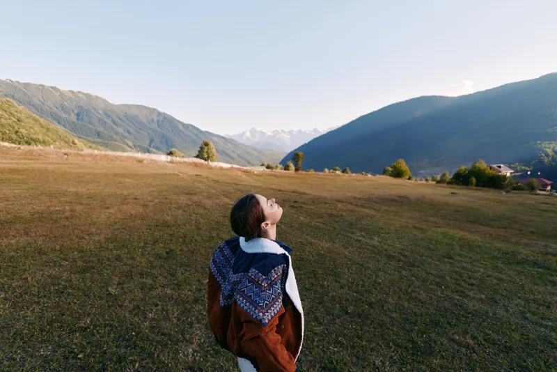 Woman in coat stands in a meadow field looking up at sky and distant mountains, serene landscape and travel concept with open space, solitude and nature scenery.