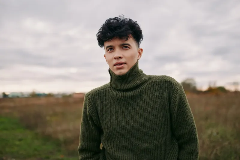 A candid portrait of a young man with curly hair wearing a thick olive sweater, conveying authenticity and believability through natural expression and pose.