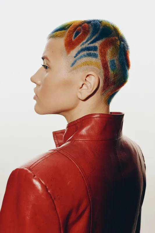 Stylish woman with short colorful hair and patterned shaved design, wearing a red leather jacket, showcasing modern fashion and creative self-expression in minimalistic background.