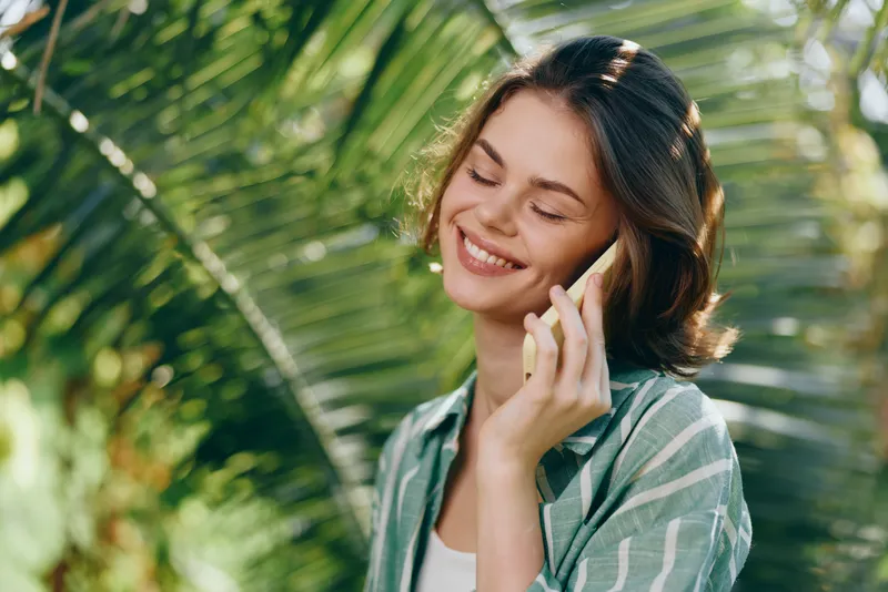 Woman chats on phone outdoors, smiling softly amid lush greenery, wearing a casual striped shirt, relaxed pose, natural light, cheerful mood, everyday moments, candid portrait in a vibrant garden