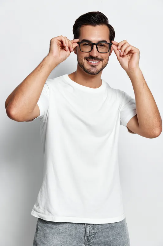 Smiling young man wearing glasses and a white t-shirt, posing playfully while adjusting his eyewear, isolated on a minimalistic background. People lifestyle concept.