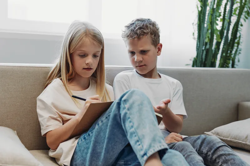 Two focused children sit on sofa indoors, girl writing on clipboard while boy explains with hand gestures. Concept of education, teamwork, learning, communication, and childhood development.