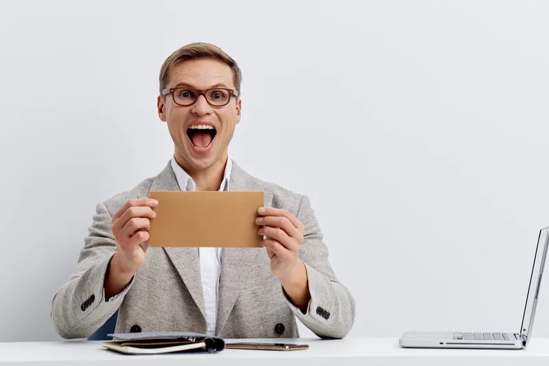 Joyful businessman holding a small cardboard box, expressing excitement or surprise, in a modern office setting with a neutral background, professional attire, and a laptop on the desk, lifestyle conc