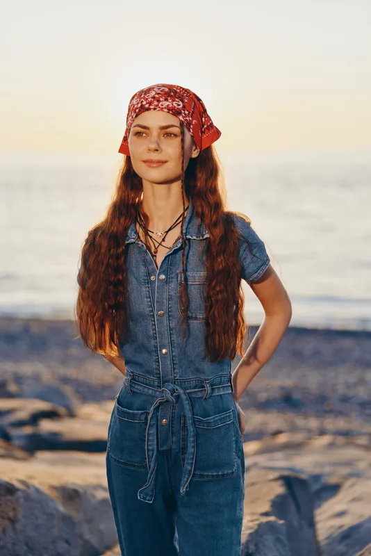 Lifestyle woman in modern boho-western denim street style stands by the sea in warm film color tones, wearing a red bandana and layered necklaces with long wavy hair.