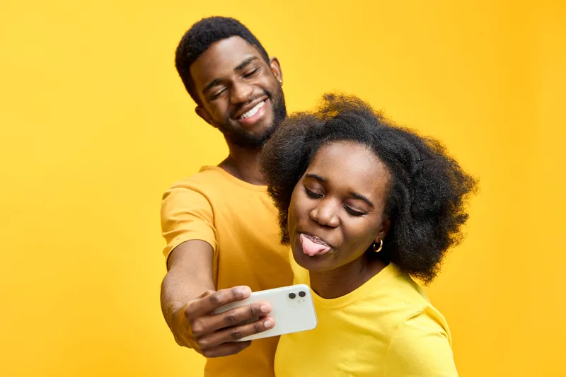 A cheerful African American couple taking a selfie against a vibrant yellow background, showcasing joyful expressions and casual yellow outfits for social media engagement
