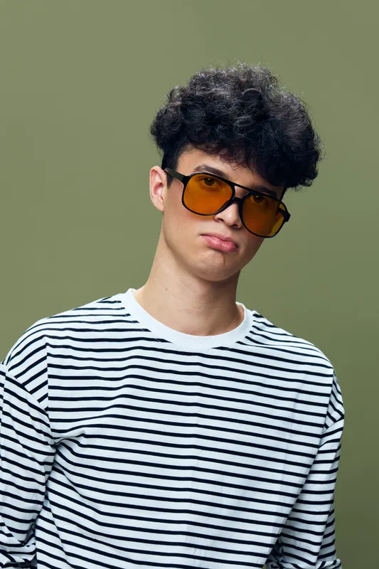 young man wearing yellow sunglasses and striped shirt poses with cool expression against green background, casual style, contemporary fashion portrait