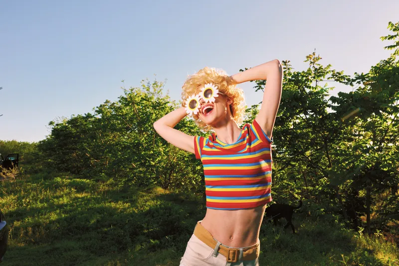Woman outdoors in a sunlit garden, wearing a striped crop top and flower sunglasses, radiating carefree retro charm, vibrant lifestyle portrait in warm tones.