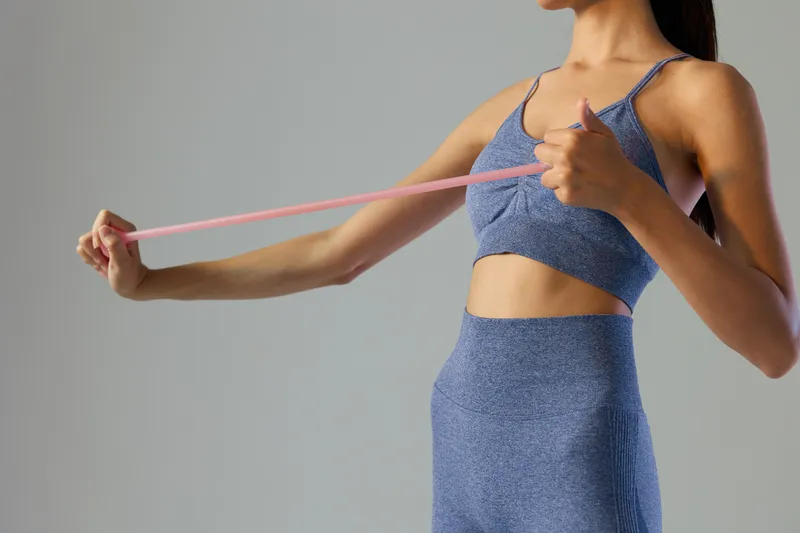 Woman sport fitness wearing blue workout outfit stretching with pink resistance band indoors, focused on exercise and body strength in a simple gray background.