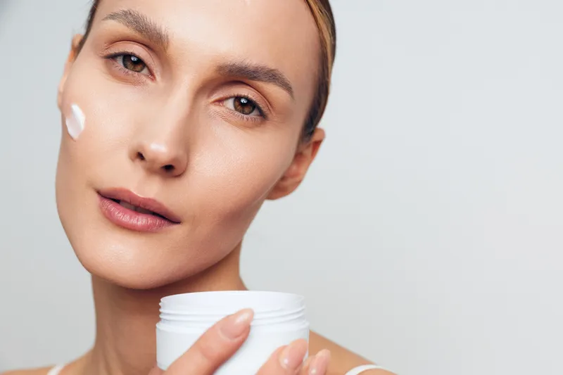 A close-up of a young woman applying face cream on her cheek with a neutral expression against a plain background. Concept of skincare, beauty, and daily routine.