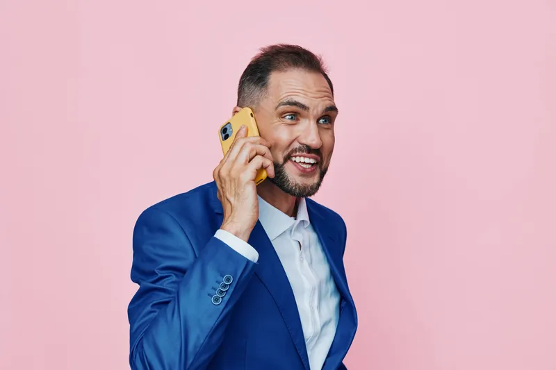 A professional man in a blue suit on an isolated pink background, speaking on a yellow phone, displaying confident posture and dynamic emotion suitable for business use.