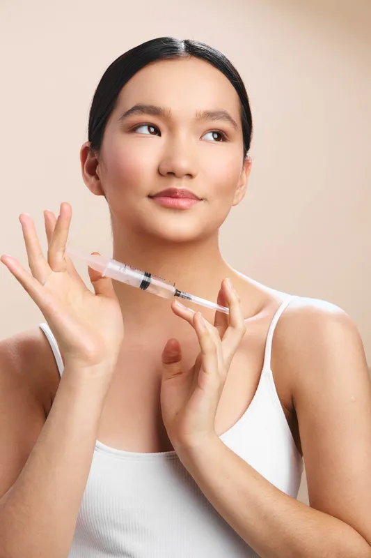 Asian woman is seen holding a syringe near her face in a clean clinical setting. She appears calm and confident, creating a sense of professional care and readiness.
