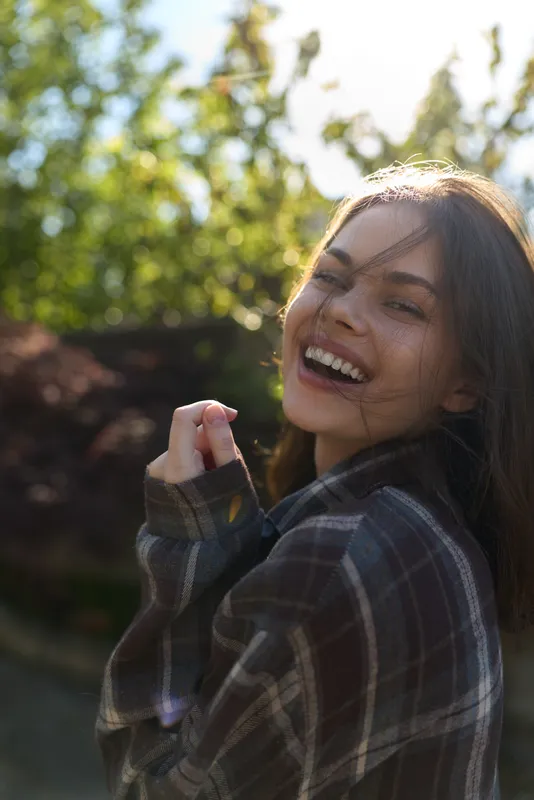 Young woman with plaid shirt smiles warmly in an outdoor setting, capturing a bright, natural moment of joy and relaxed confidence