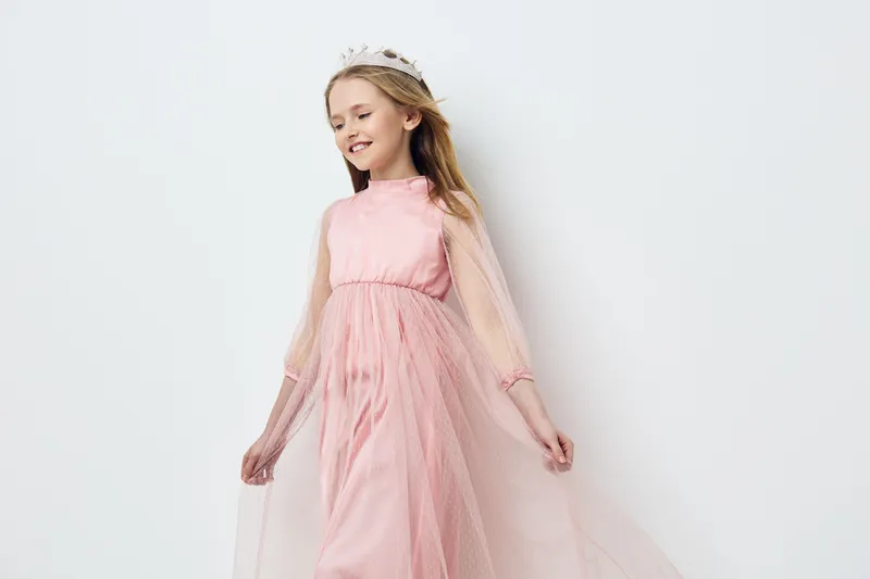 A joyful young girl in a pink dress twirls gracefully with a crown, showcasing elegance and innocence in a bright, minimalist setting