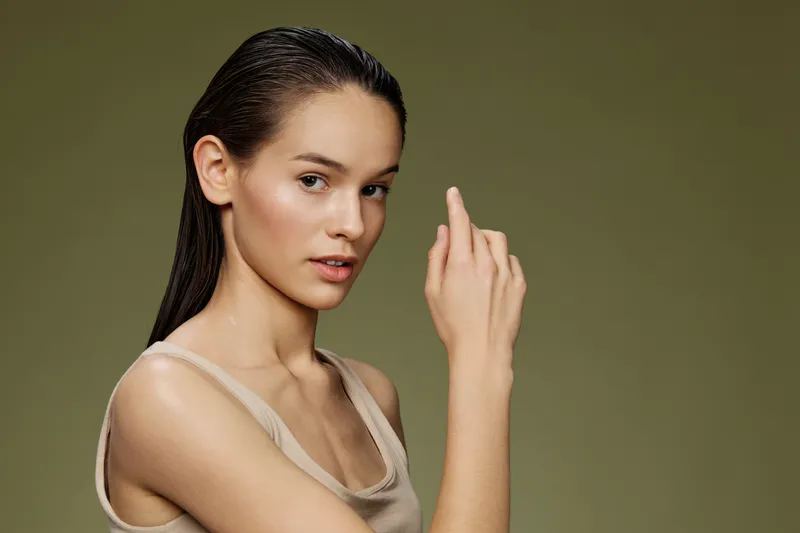 A confident young woman with long hair poses against a muted green background, showcasing healthy skin and a neutral outfit, embodying elegance and strength