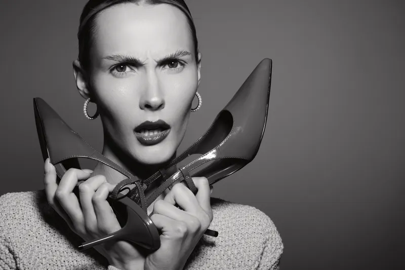 Young white woman with light skin holds high heel shoes near face with confused expression in studio. Concept of fashion, style, and emotions.