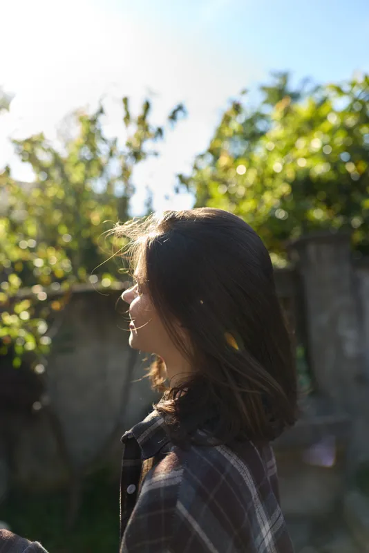 Young woman with brunette hair stands in a sunlit backyard, gazing to the side as soft light filters through leaves, wearing a plaid shirt and a relaxed casual mood