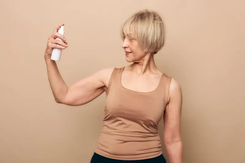 Mature woman with short blonde hair in beige tank top sprays facial mist on her face, isolated on neutral background. Concept of skincare, hydration, and self-care for older adults.