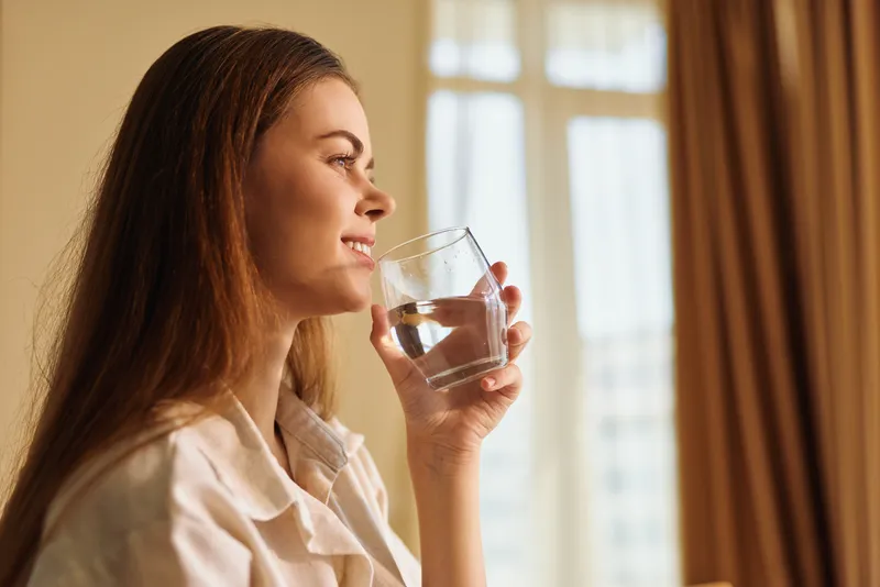 Woman with glass of water drinking and smiling by window in morning sunlight for hydration and healthy lifestyle, portrait at home, relaxed routine, wellness and calm domestic scene.