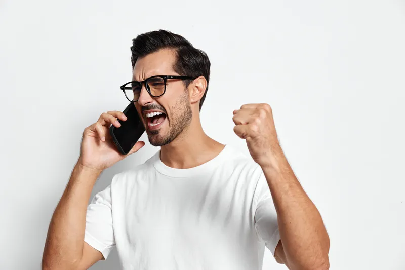 Angry young man wearing glasses, shouting on smartphone, expressing frustration with raised fist, isolated on plain background. Concept of communication and emotions.
