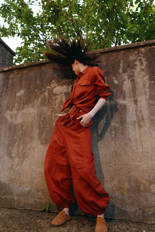 A woman in a bold red jumpsuit dances beside a concrete wall, hair in motion. The moment captures dynamic street style, confidence and carefree movement