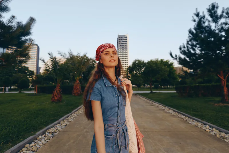 Lifestyle woman in modern boho-western denim street style stands confidently on an urban path with greenery and skyscrapers, captured in warm film color tones.