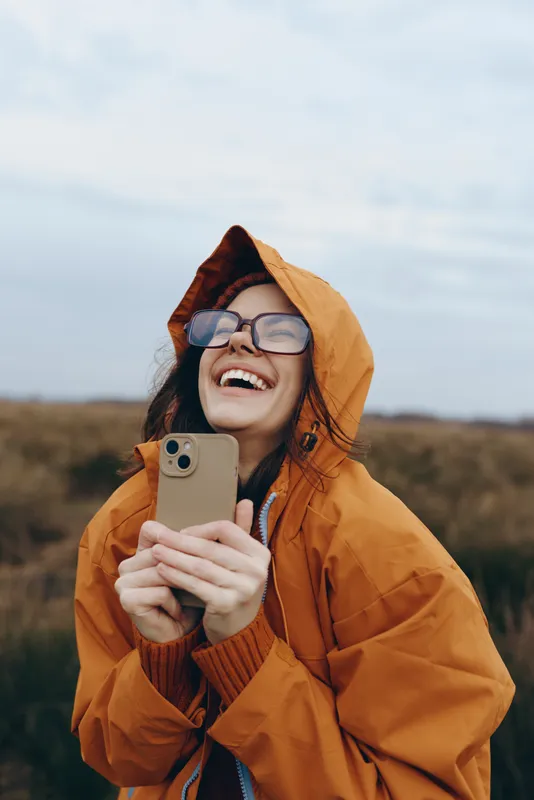 Woman lifestyle in autumn outdoors, laughing smiling in orange hood coat, wearing glasses, holding smartphone in hands, natural landscape background, warm casual mood, fresh air today